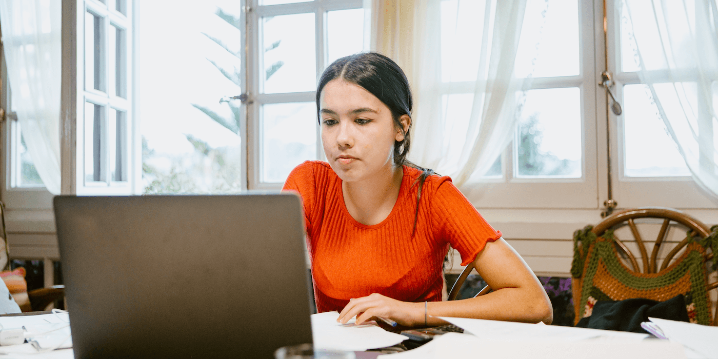 college student sitting at home at a table with papers around them and typing on a laptop
