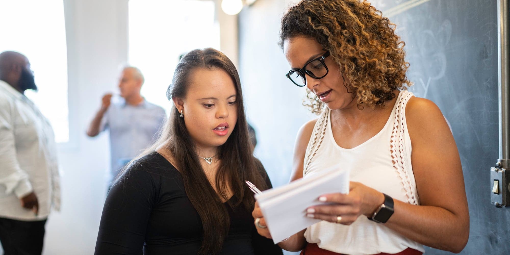 Two people standing in front of a blackboard looking at the same notebook, with two other people out of focus in background talking