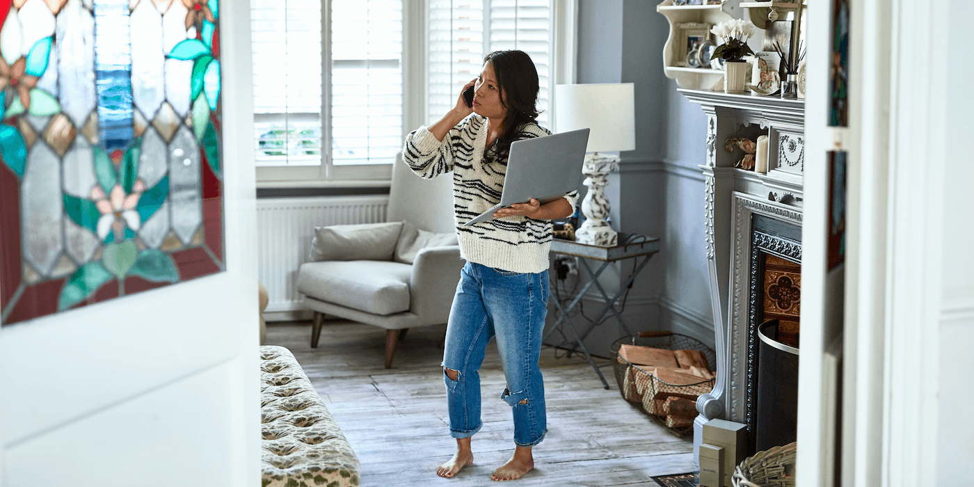 person standing at home holding an open laptop and speaking on a cell phone
