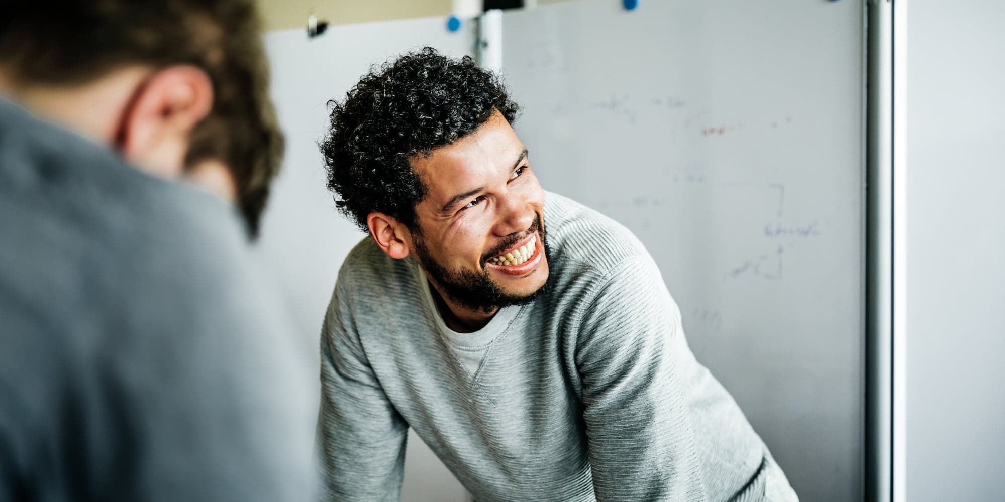 person leaning forward on table or similar, smiling and looking off to the side. Person in foreground facing away from camera and blurry